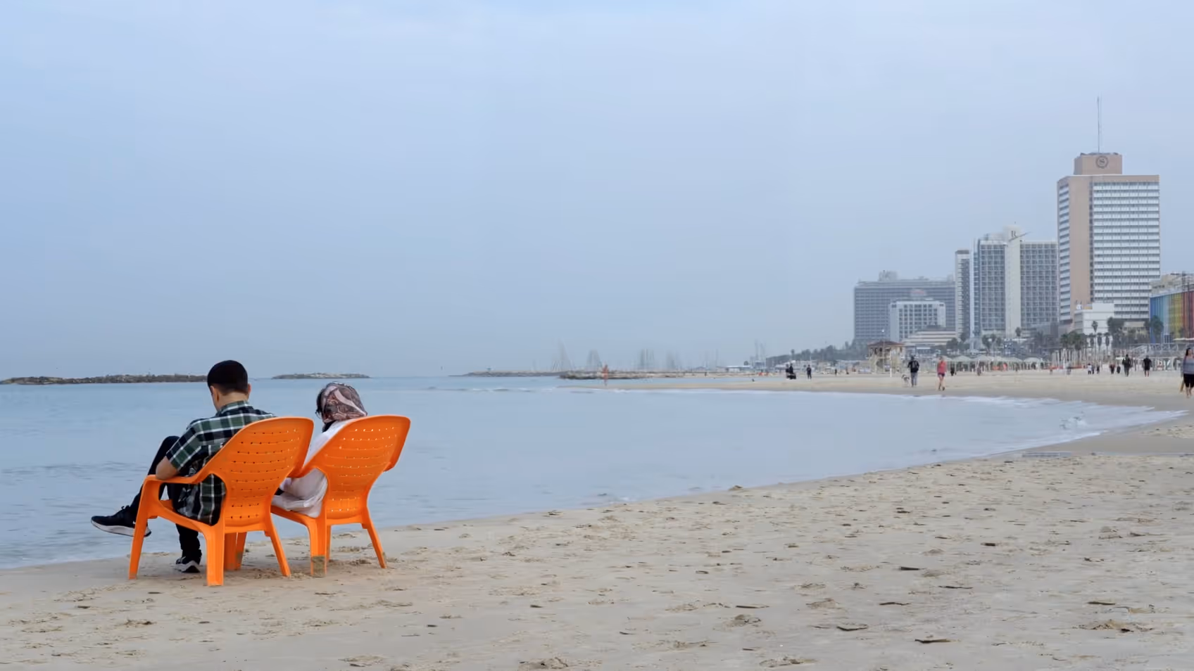 Two people seated on orange chairs on a Tel Aviv beach, with the city's seafront hotels in the background, from the documentary series Secularism
