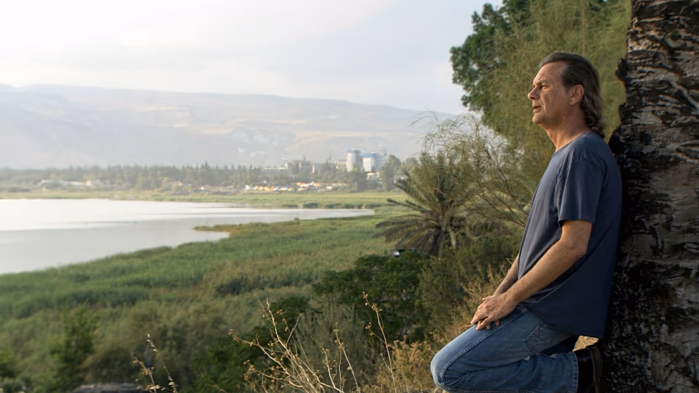 A man leaning against a tree gazing over a lush green valley and lake landscape, from the documentary series The Kibbutz